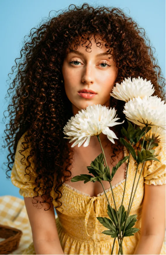 A woman with curly hair holding white chrysanthemum flowers against a sky blue backdrop.